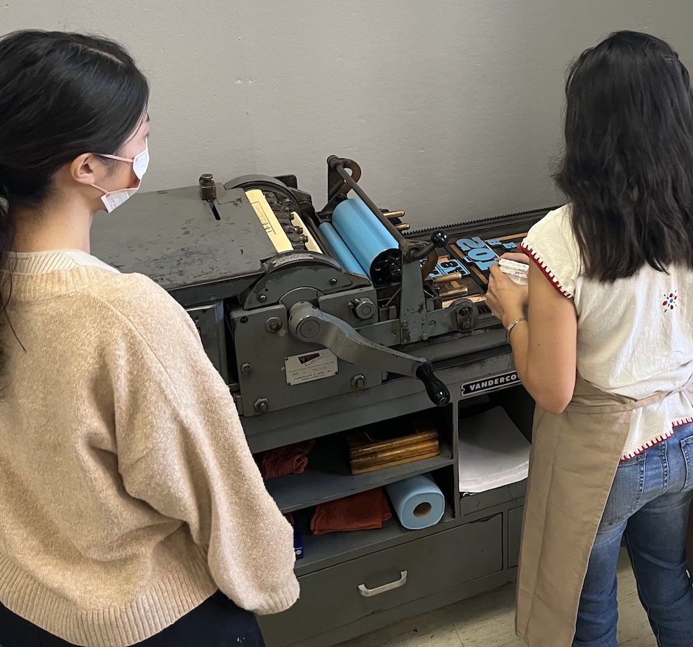 Photograph of two people with long black hair facing a proofing press.